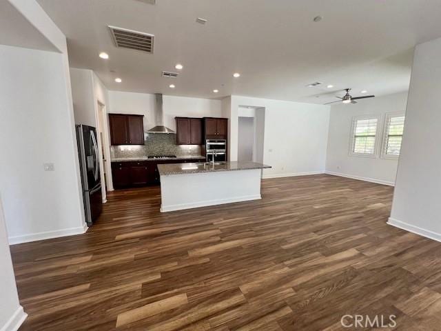 kitchen with black refrigerator, a kitchen island with sink, dark wood-type flooring, and wall chimney range hood