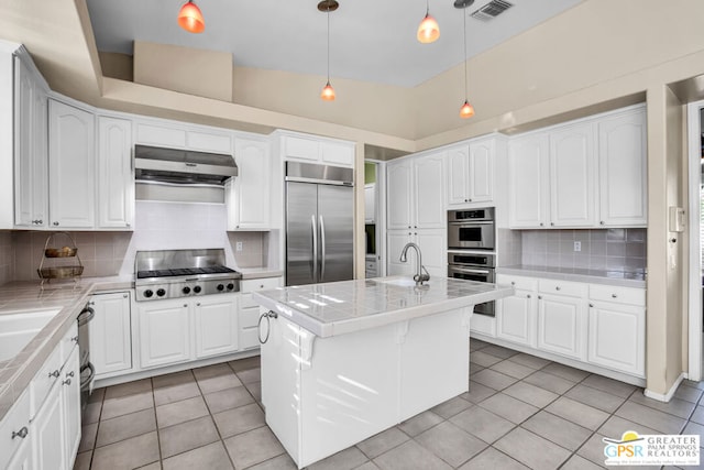 kitchen featuring appliances with stainless steel finishes, backsplash, a kitchen island with sink, exhaust hood, and hanging light fixtures