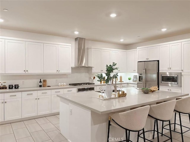 kitchen featuring white cabinetry, a center island with sink, appliances with stainless steel finishes, light tile patterned flooring, and wall chimney range hood