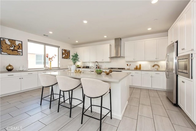 kitchen with an island with sink, white cabinets, appliances with stainless steel finishes, and wall chimney range hood