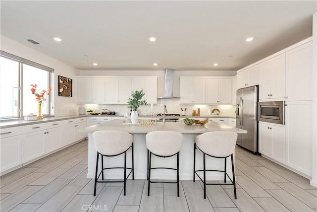 kitchen featuring stainless steel appliances, wall chimney exhaust hood, white cabinets, and a center island with sink