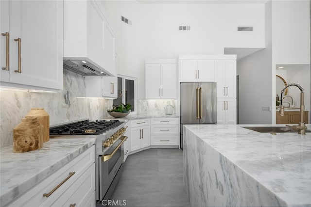 kitchen with high end appliances, white cabinetry, light stone counters, and custom range hood