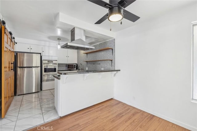 kitchen with a barn door, appliances with stainless steel finishes, white cabinetry, island range hood, and a peninsula