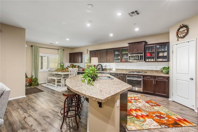 kitchen featuring appliances with stainless steel finishes, a kitchen bar, a kitchen island with sink, dark brown cabinetry, and light wood-type flooring