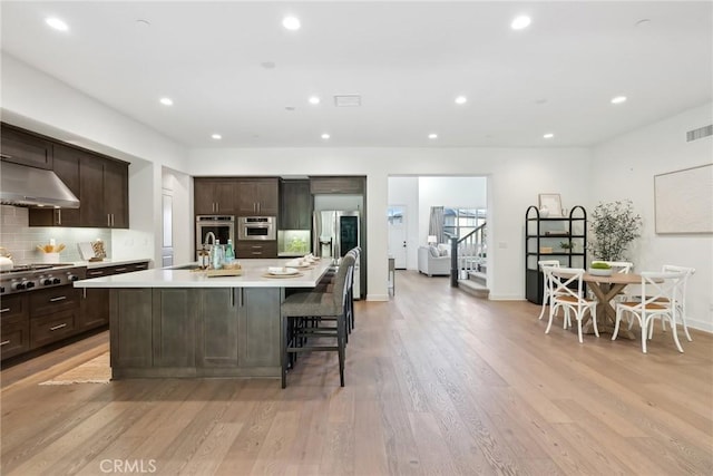 kitchen with extractor fan, a kitchen breakfast bar, a kitchen island with sink, dark brown cabinets, and light wood-type flooring