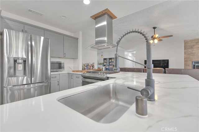 kitchen with gray cabinets, island exhaust hood, ceiling fan, stainless steel appliances, and light stone countertops