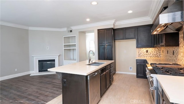 kitchen featuring wall chimney range hood, sink, built in features, an island with sink, and appliances with stainless steel finishes