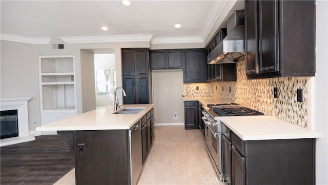 kitchen with wall chimney range hood, double oven range, sink, a kitchen island with sink, and ornamental molding