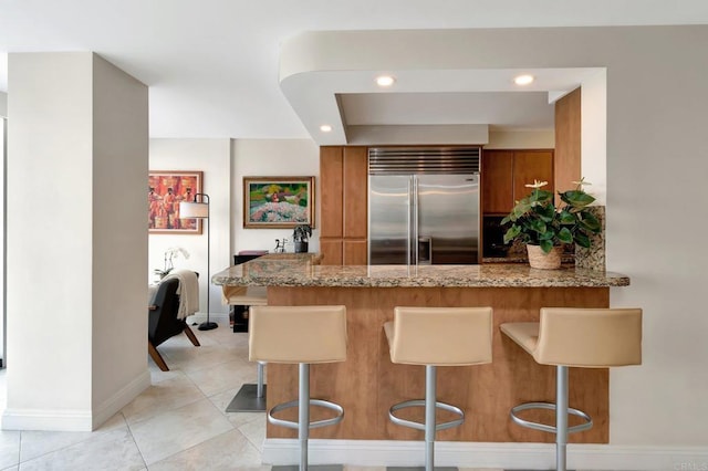 kitchen with a peninsula, light stone counters, brown cabinetry, and stainless steel built in fridge