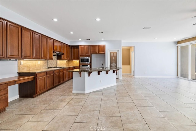 kitchen featuring under cabinet range hood, a breakfast bar, decorative backsplash, dark stone countertops, and stainless steel appliances