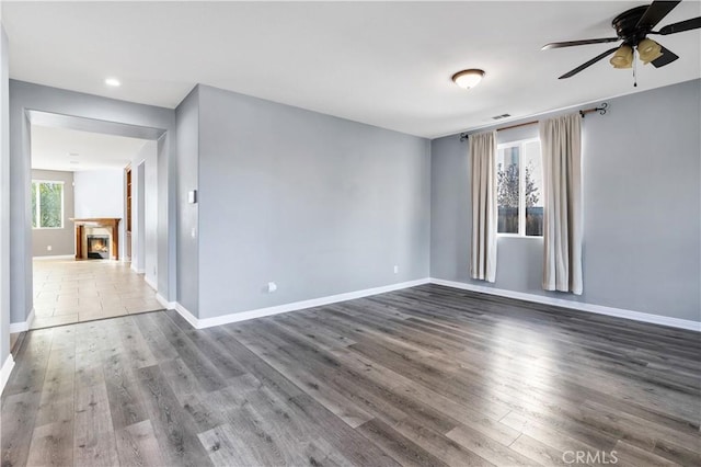 spare room featuring ceiling fan, baseboards, a warm lit fireplace, and wood finished floors