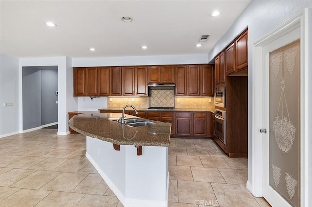 kitchen with an island with sink, a sink, dark stone countertops, appliances with stainless steel finishes, and decorative backsplash