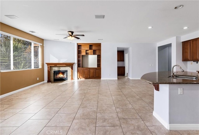 kitchen featuring light tile patterned floors, baseboards, dark stone counters, a warm lit fireplace, and a sink