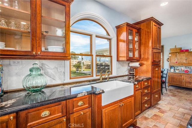 kitchen featuring glass insert cabinets, dark stone counters, a sink, stone finish flooring, and backsplash