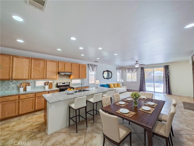 kitchen with a breakfast bar, stainless steel range oven, light stone counters, a center island with sink, and backsplash