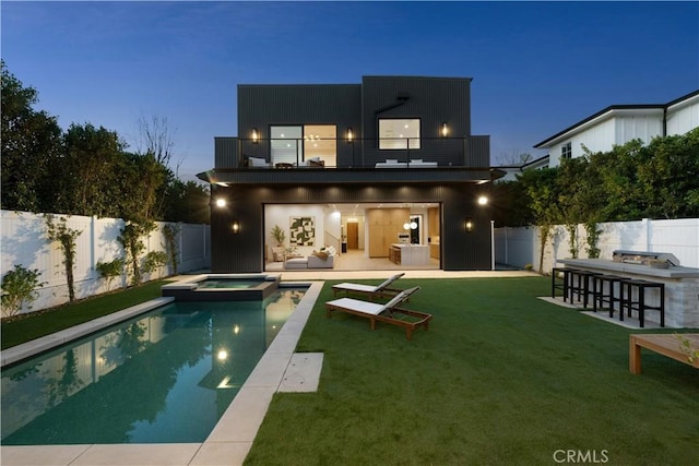 back house at dusk with a balcony, exterior kitchen, a swimming pool with hot tub, a yard, and an outdoor bar