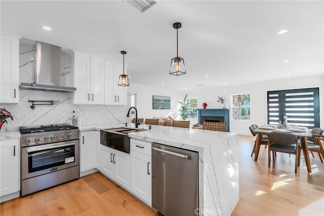 kitchen featuring kitchen peninsula, stainless steel appliances, wall chimney exhaust hood, white cabinets, and sink