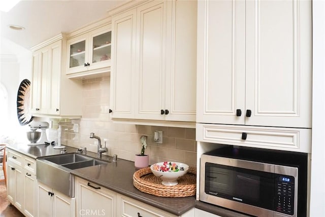 kitchen featuring tasteful backsplash, dark countertops, stainless steel microwave, glass insert cabinets, and a sink