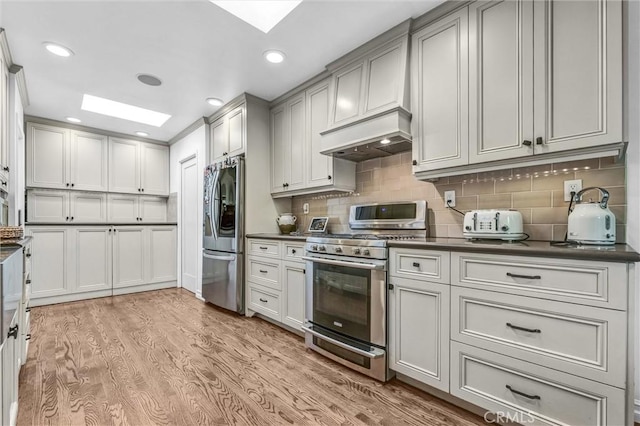 kitchen with a skylight, stainless steel appliances, dark countertops, custom range hood, and light wood-style flooring