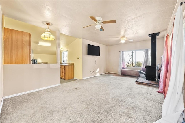 unfurnished living room with ceiling fan, a textured ceiling, light colored carpet, baseboards, and a wood stove