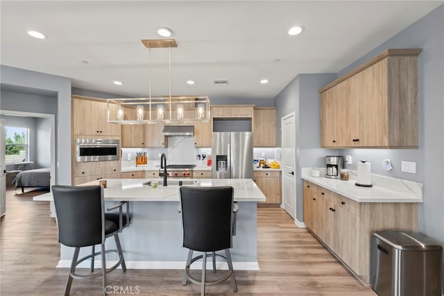 kitchen with appliances with stainless steel finishes, light brown cabinetry, hanging light fixtures, and wall chimney range hood