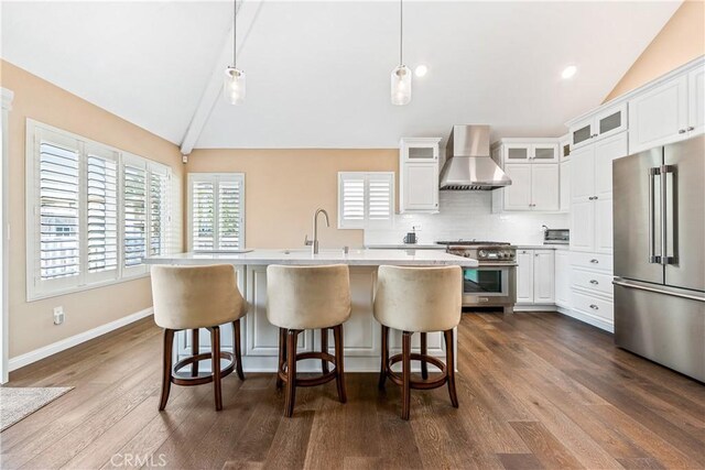 kitchen featuring premium appliances, white cabinets, decorative light fixtures, wall chimney range hood, and a center island with sink