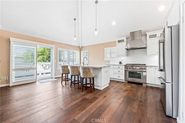 kitchen with white cabinets, appliances with stainless steel finishes, wall chimney exhaust hood, hanging light fixtures, and a center island with sink