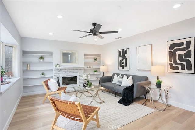 living room with a brick fireplace, built in features, ceiling fan, and light wood-type flooring