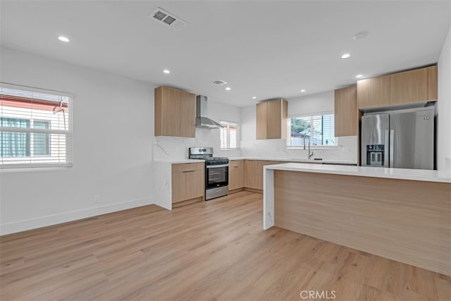 kitchen featuring wall chimney range hood, stainless steel appliances, light brown cabinetry, sink, and light hardwood / wood-style flooring