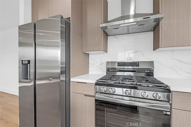 kitchen featuring appliances with stainless steel finishes, wall chimney range hood, tasteful backsplash, light brown cabinetry, and light wood-type flooring