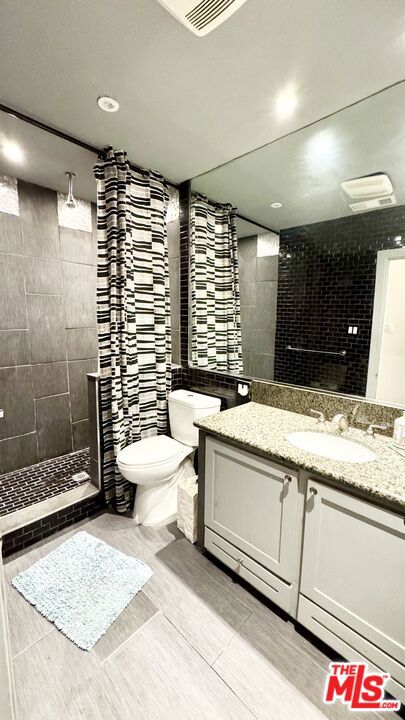 bathroom featuring vanity, tile patterned flooring, toilet, tiled shower, and tile walls