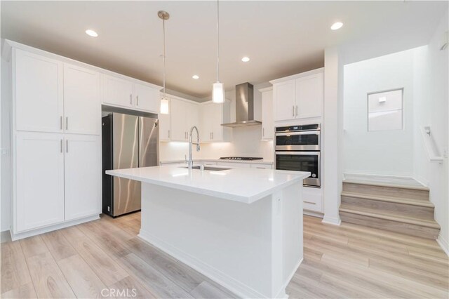 kitchen with decorative light fixtures, sink, a kitchen island with sink, appliances with stainless steel finishes, and wall chimney exhaust hood