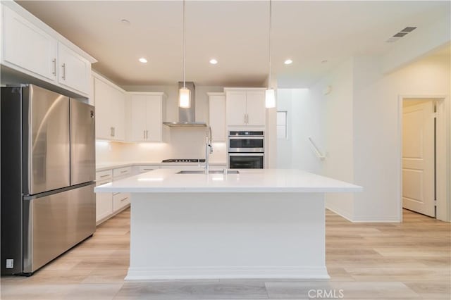 kitchen with pendant lighting, white cabinets, appliances with stainless steel finishes, wall chimney exhaust hood, and a center island with sink