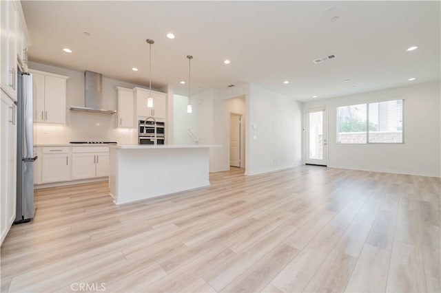 kitchen with white cabinets, wall chimney range hood, an island with sink, hanging light fixtures, and stainless steel fridge