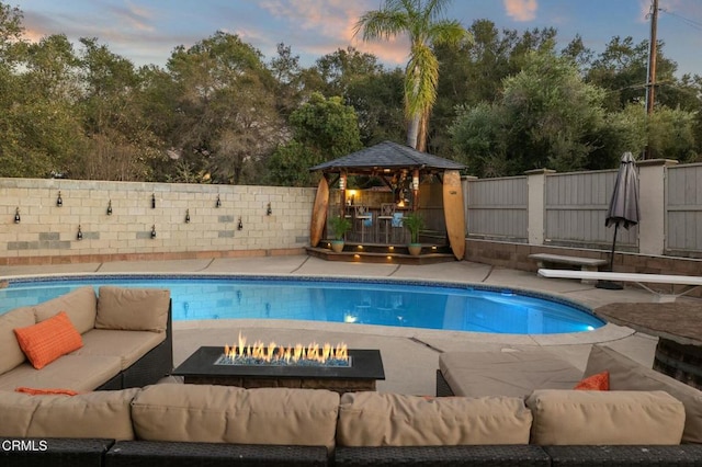 pool at dusk with a fenced in pool, a fenced backyard, a diving board, an outdoor living space with a fire pit, and a gazebo