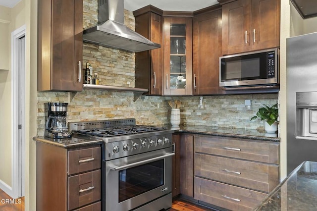 kitchen featuring dark stone counters, wall chimney range hood, tasteful backsplash, glass insert cabinets, and appliances with stainless steel finishes
