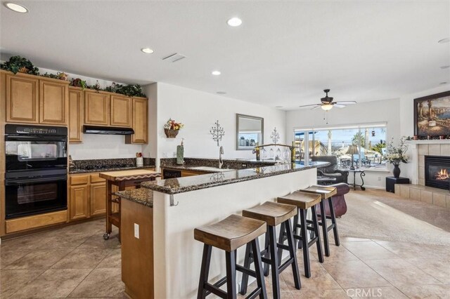 kitchen with double oven, a fireplace, dark stone countertops, a kitchen breakfast bar, and ceiling fan