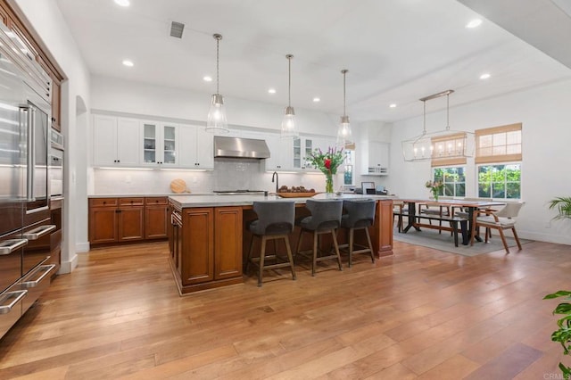 kitchen with wall chimney range hood, light hardwood / wood-style floors, hanging light fixtures, an island with sink, and white cabinets