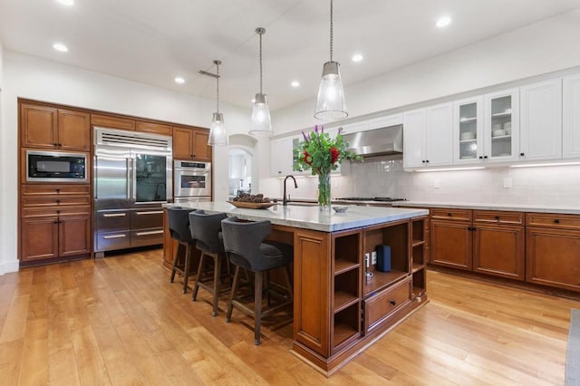 kitchen featuring an island with sink, backsplash, wall chimney range hood, white cabinets, and built in appliances