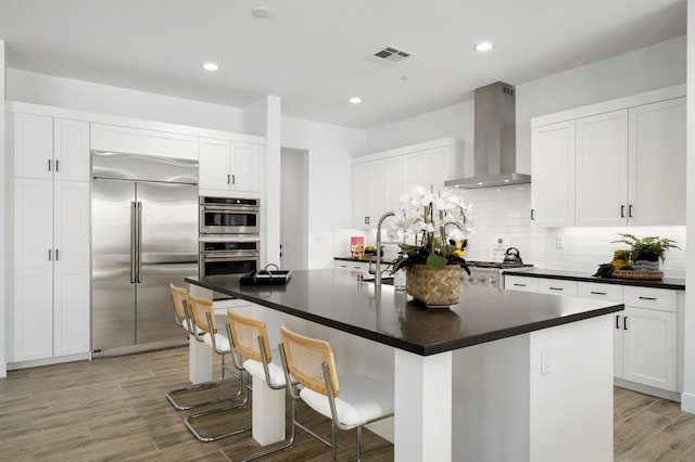 kitchen with a kitchen bar, stainless steel appliances, a kitchen island with sink, wall chimney exhaust hood, and sink