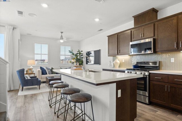 kitchen featuring light wood-type flooring, stainless steel appliances, dark brown cabinetry, and a center island with sink