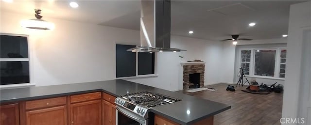 kitchen featuring ceiling fan, stainless steel range with gas stovetop, island range hood, a stone fireplace, and kitchen peninsula