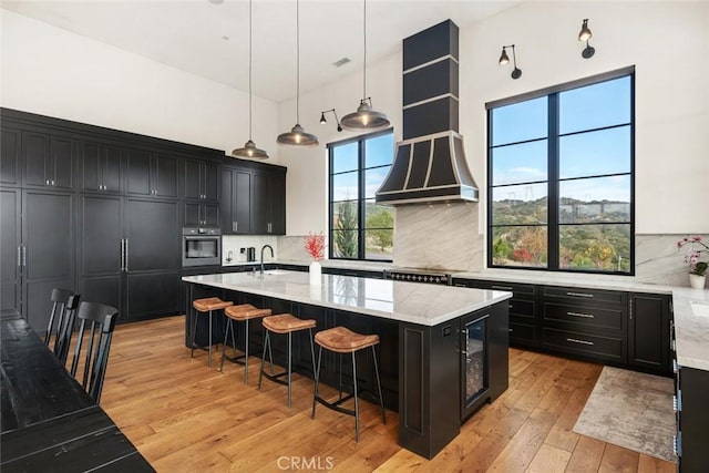 kitchen with an island with sink, backsplash, oven, light stone countertops, and pendant lighting