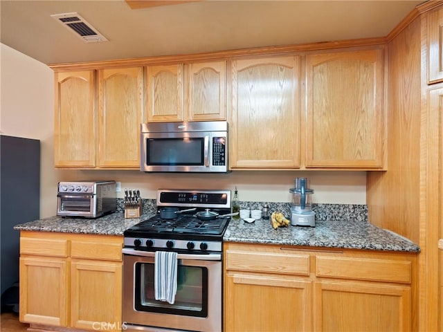 kitchen with light brown cabinetry, appliances with stainless steel finishes, and dark stone counters