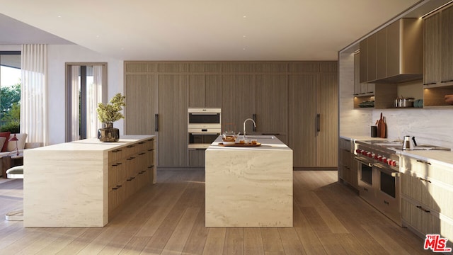 kitchen with double oven range, a center island with sink, double wall oven, light hardwood / wood-style flooring, and sink