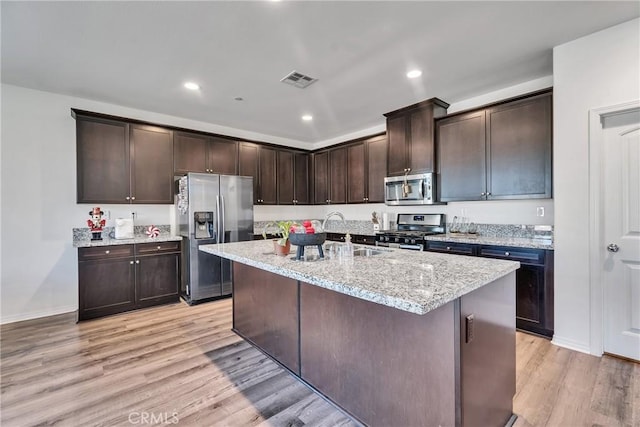 kitchen featuring light stone countertops, stainless steel appliances, an island with sink, light hardwood / wood-style flooring, and dark brown cabinets