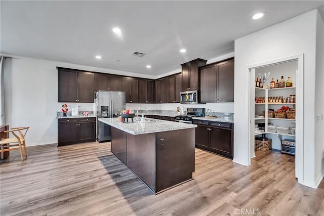 kitchen with a center island with sink, appliances with stainless steel finishes, dark brown cabinets, light stone countertops, and light hardwood / wood-style flooring