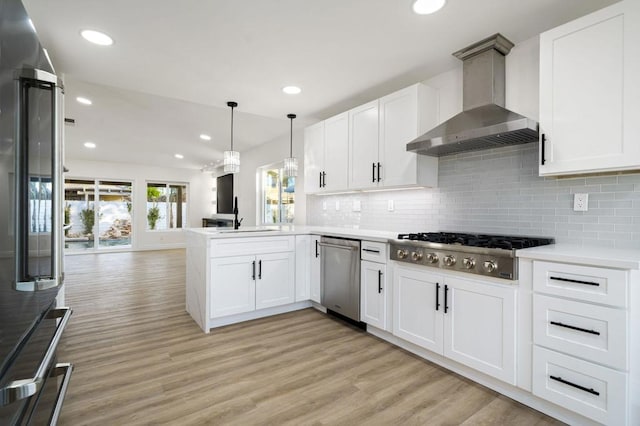 kitchen featuring white cabinetry, kitchen peninsula, stainless steel appliances, hanging light fixtures, and wall chimney range hood