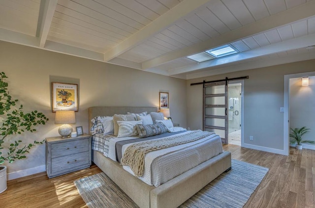 bedroom with a skylight, a barn door, light hardwood / wood-style floors, ensuite bath, and beam ceiling