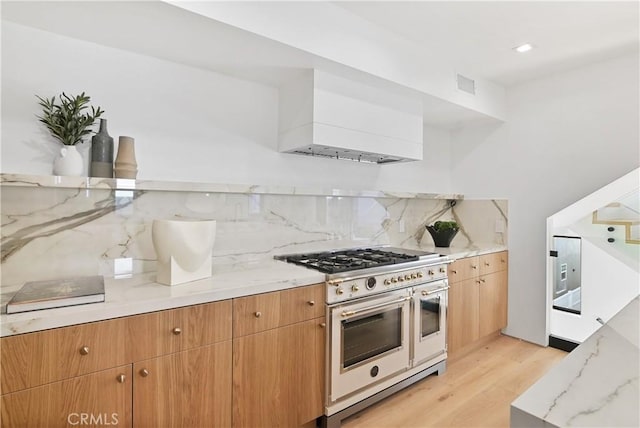 kitchen with double oven range, custom exhaust hood, tasteful backsplash, light stone countertops, and light hardwood / wood-style flooring